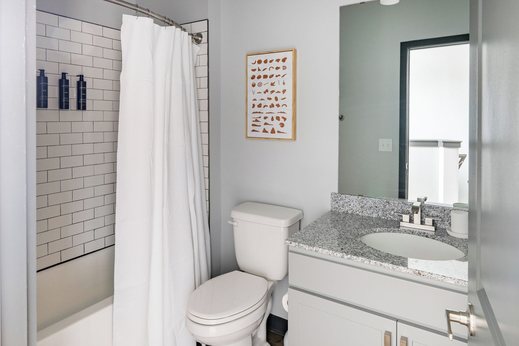 Une salle de bain moderne avec des toilettes blanches, une douche avec un rideau blanc, un lavabo sur un plan de travail en granit et un tableau encadré au mur.