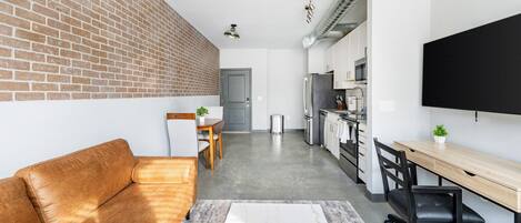 A modern apartment with a brick wall, a sofa, a dining area, a kitchen, and a TV.