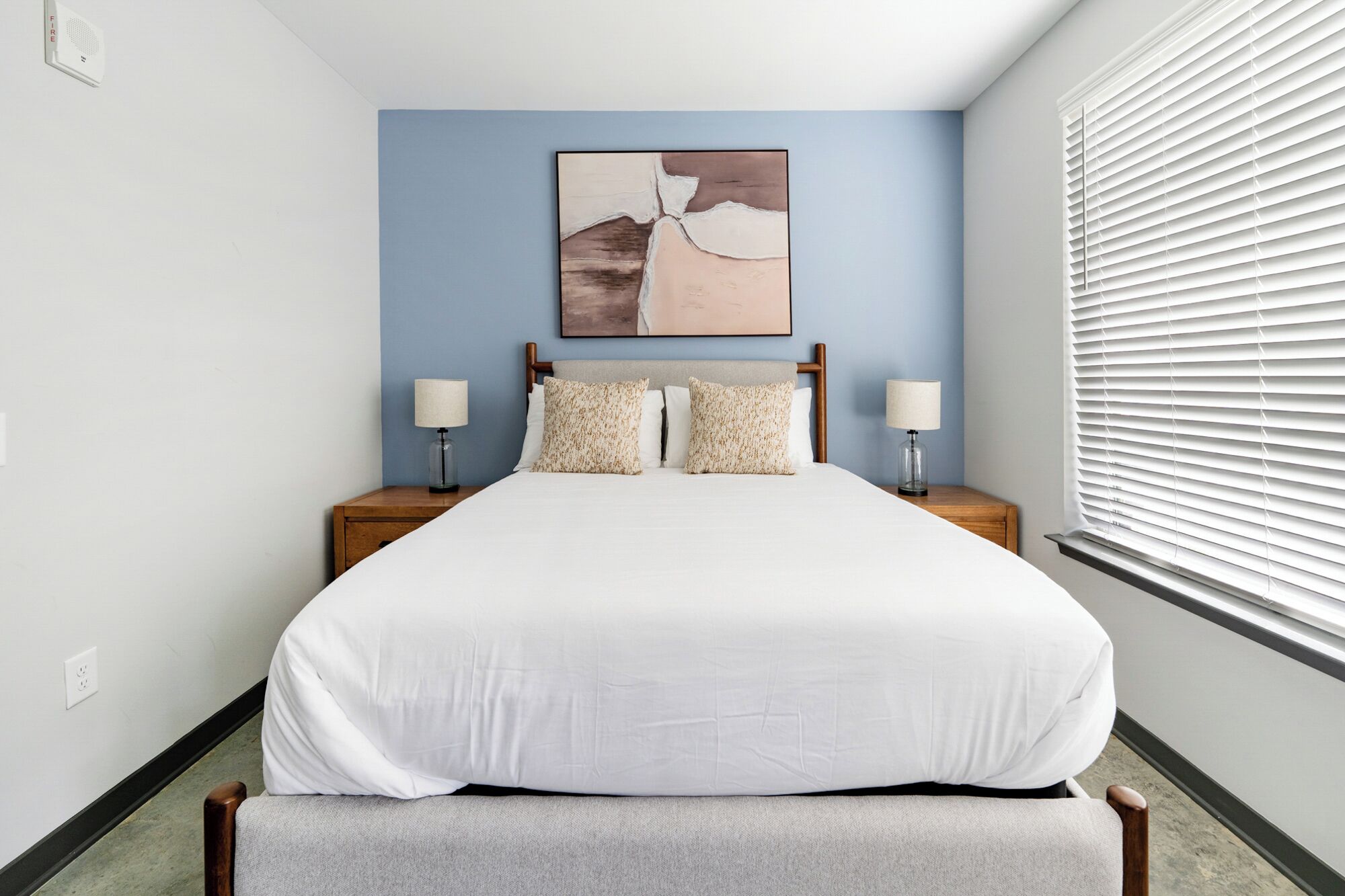 A neatly arranged bedroom with a large bed, two bedside tables with lamps, a headboard with a painting, and a window with blinds.