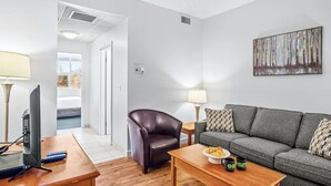 Ett modernt vardagsrum med en grå soffa, en brun läderfåtölj, ett träbord med en tallrik mat och två muggar, en tv på ett träställ och en tavla på väggen.