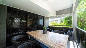 Ein Balkon mit einem Holztisch, Korbstühlen, einem Blumentopf und Blick auf Grünflächen sowie ein Gebäude.