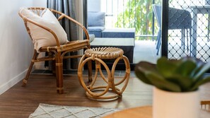 Ein Korbstuhl mit Kissen und ein runder Hocker in einem Zimmer mit Holzboden, einem weißen Teppich und einer Topfpflanze.