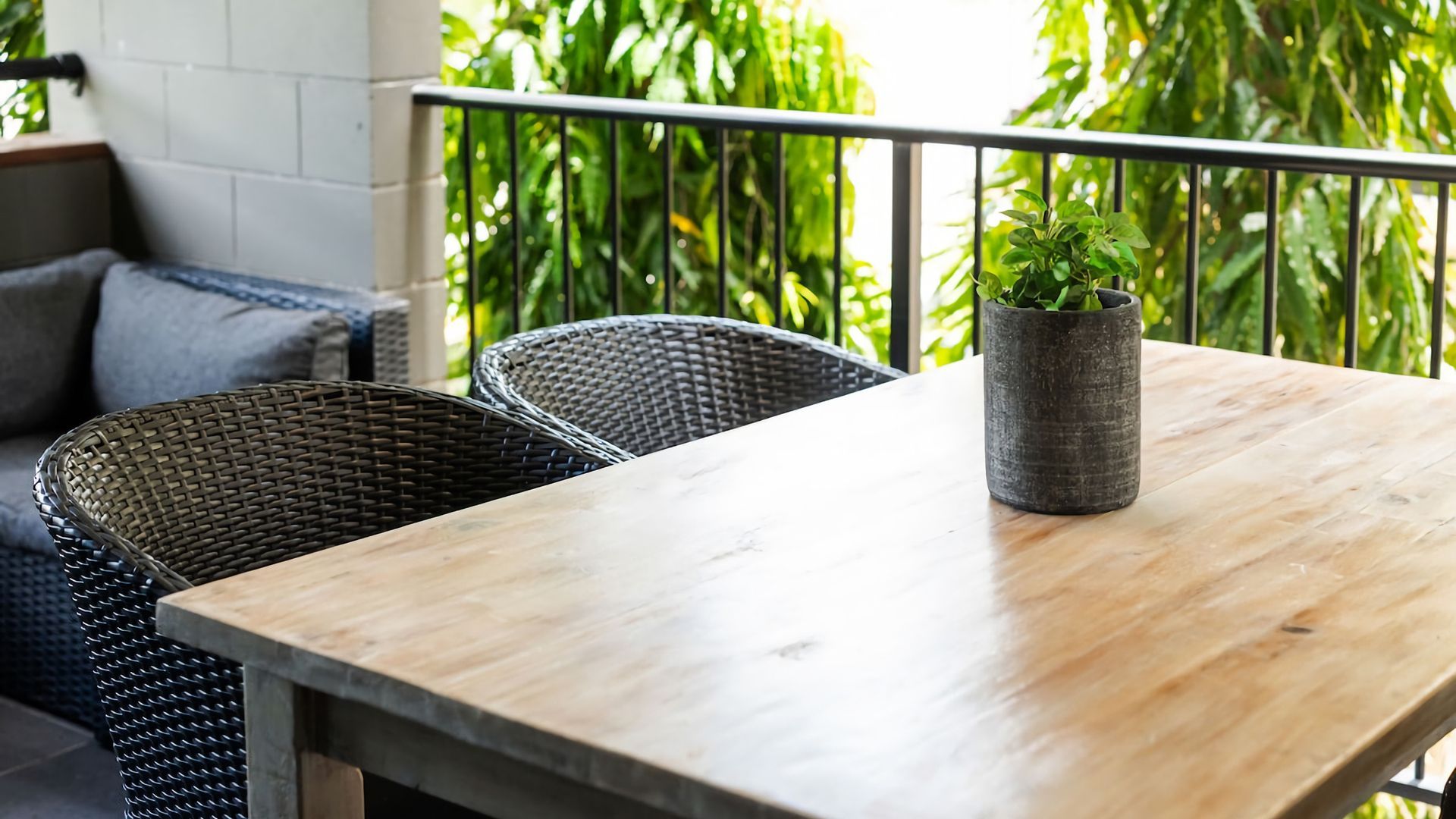 A wooden table with a potted plant, wicker chairs, and a balcony with greenery.