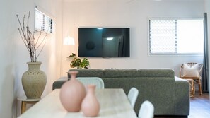 Ein Wohnzimmer mit grauem Sofa, einem flachbildfernseher an der Wand, einem großen Fenster mit Jalousien und einem kleinen Tisch mit dekorativen Vasen.