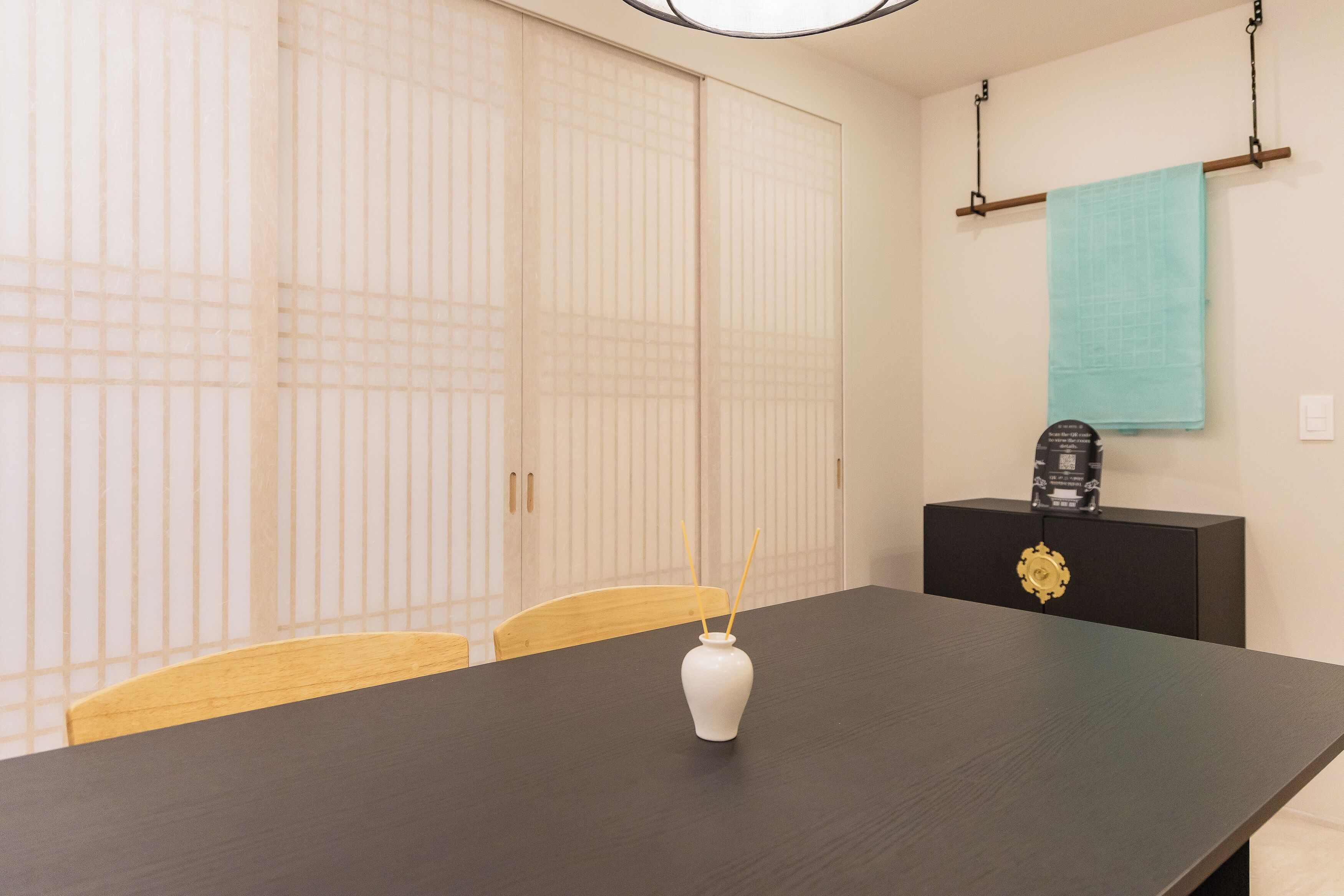A room with a dark wooden table, a white vase, a black cabinet with a gold ornament, and a hanging blue towel.
