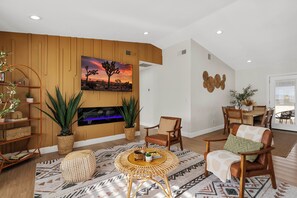 A living room with a fireplace, wooden panel walls, a dining area, and a large window.