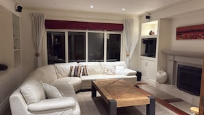 Een moderne woonkamer met een hoekbank, een houten salontafel, een open haard en grote ramen met gordijnen.