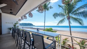 A terrace with a view of the beach and ocean, featuring a long table and chairs, potted plants, and hanging wicker lamps. - Ozeano Jaco (Jacó)