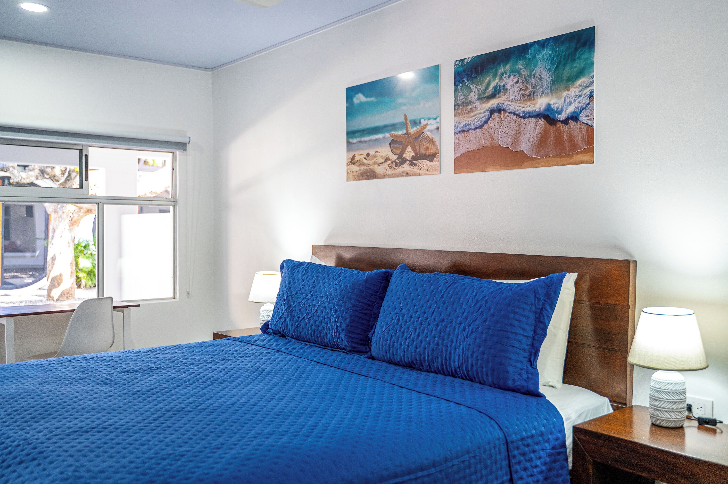 Habitación de hotel con una cama grande, cabecero de madera, dos lámparas fijadas a la pared y dos cuadros con motivos marinos.