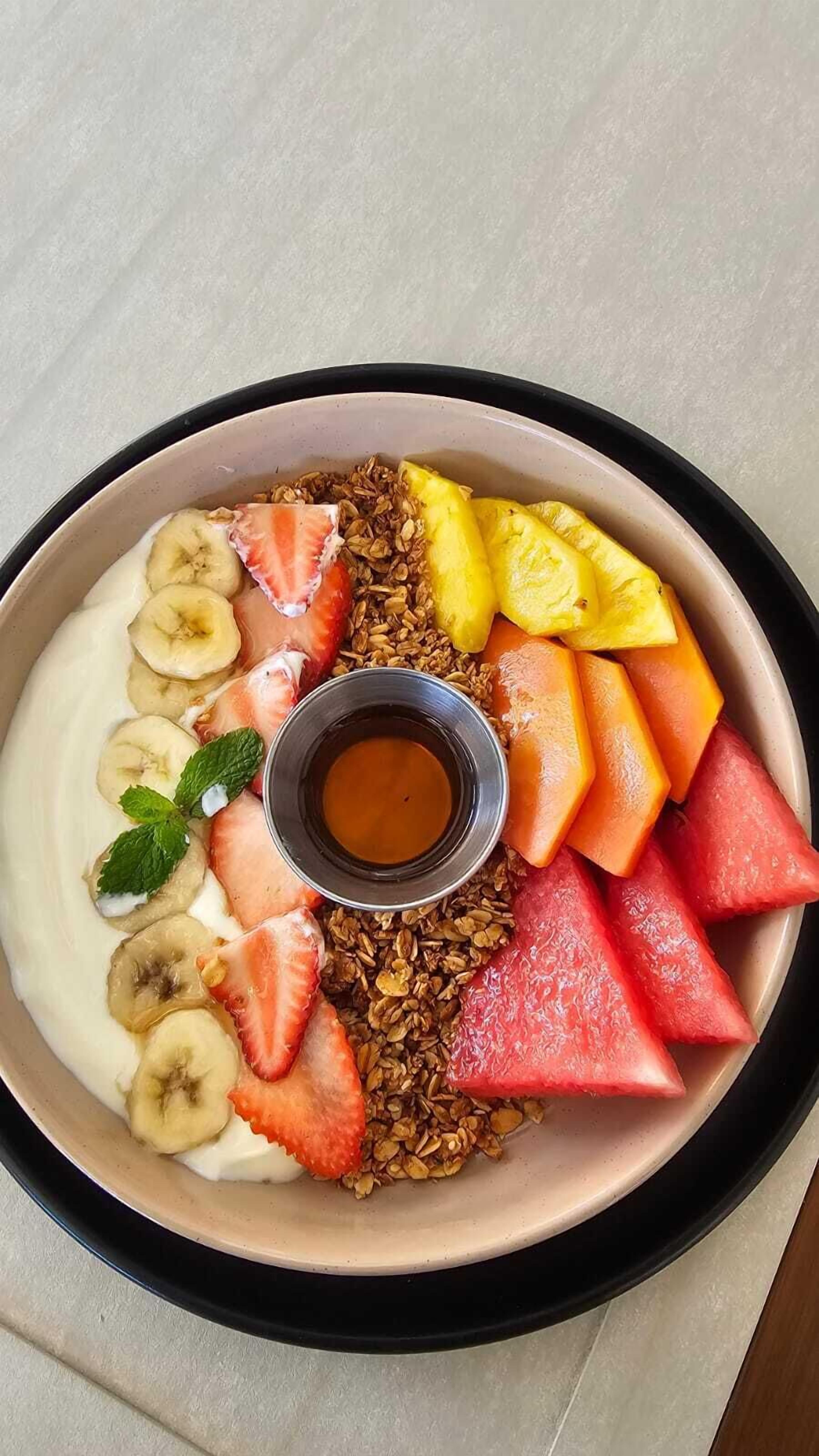 A bowl of yogurt with granola, strawberries, bananas, watermelon, and pineapple slices, accompanied by a small bowl of syrup.