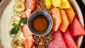 Un bol de iogurt amb granola, maduixes, plàtans, taronges i talls d'anana, acompanyat d'un bol petit de xarop.