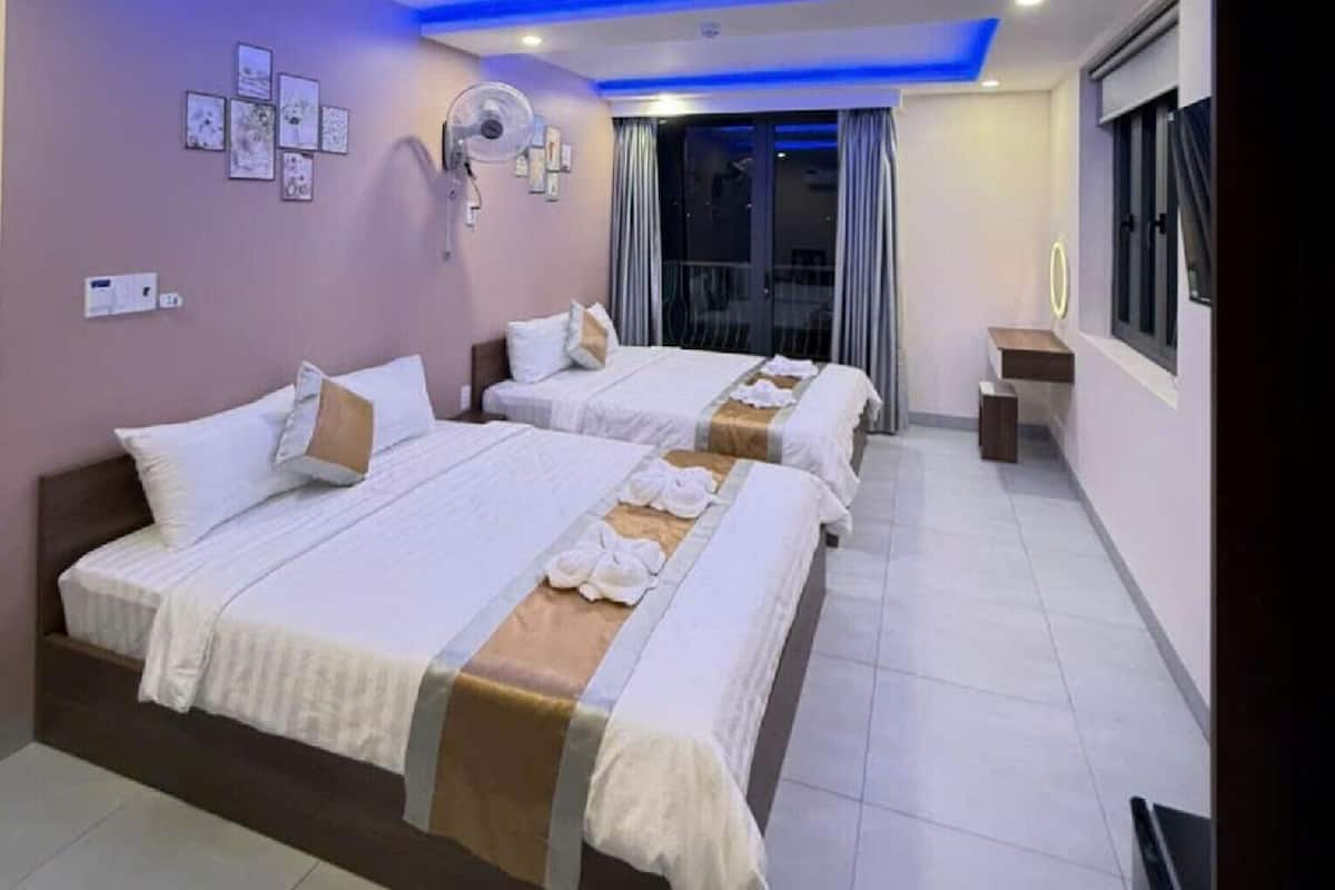 A hotel room with two beds, a ceiling fan, a small table, and a window with curtains.