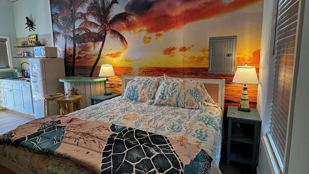 Un dormitorio con cama, mesitas de noche, ventilador de techo, un mural de atardecer y una ventana con persianas.