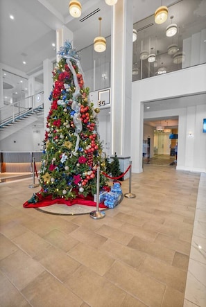 Un grand sapin de Noël décoré de guirlandes et d’illuminations, installé à l’intérieur, entouré d’un tapis rouge et d’une rambarde en verre.