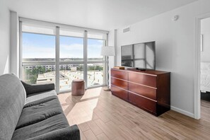 A modern hotel room with a large window, a sofa, a wooden cabinet, and a flat-screen TV. - Cozy Apartment in the Heart of Fort Myers (Fort Myers)