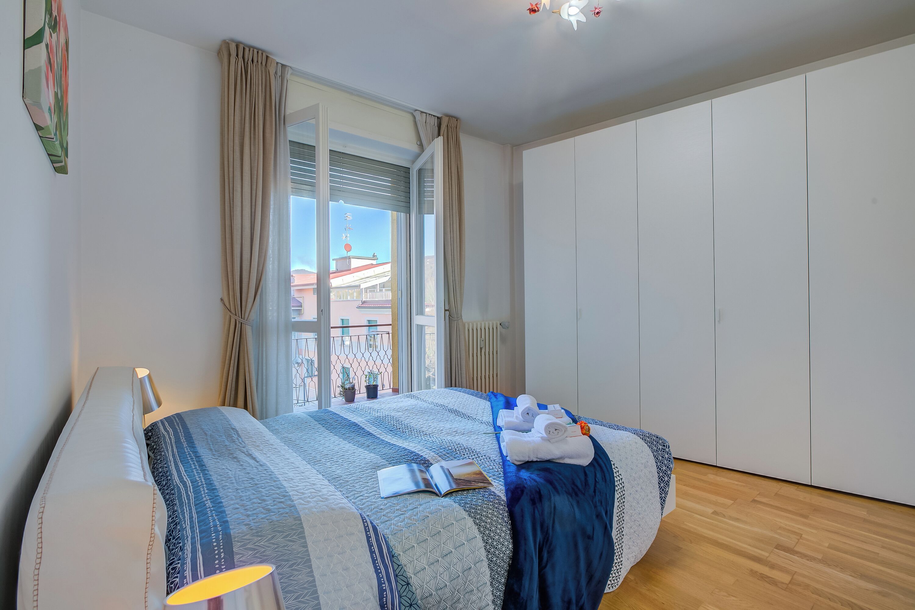 A hotel room with a bed, a window with curtains, a view of buildings, and a wardrobe.