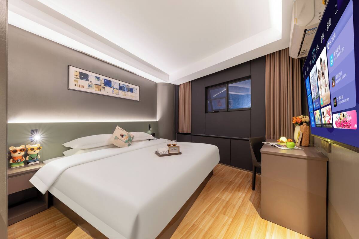 Una habitación de hotel moderna con una cama bien hecha, una mesita de noche con una lámpara, un televisor pequeño y un escritorio con un jarrón y flores.