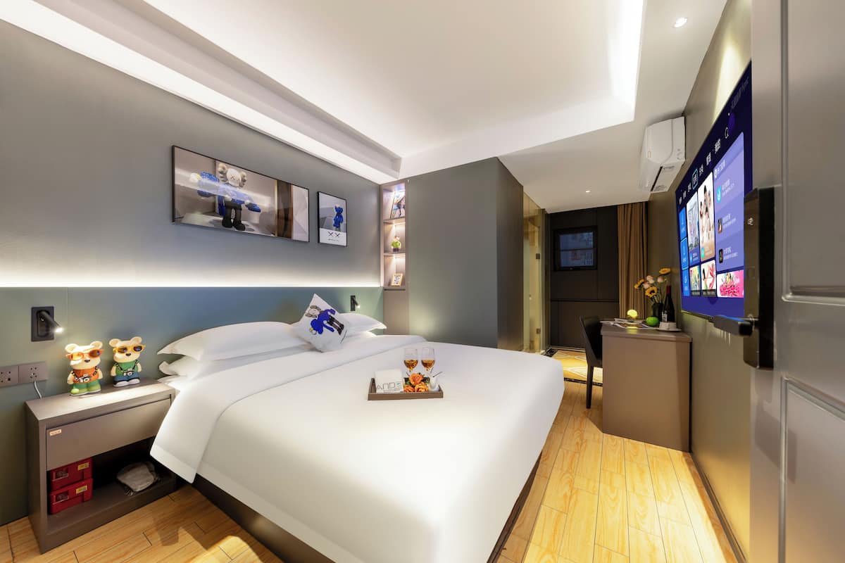 Una habitación de hotel moderna con una cama bien hecha, una mesita de noche con lámpara, un televisor montado en la pared y una pequeña zona de escritorio.