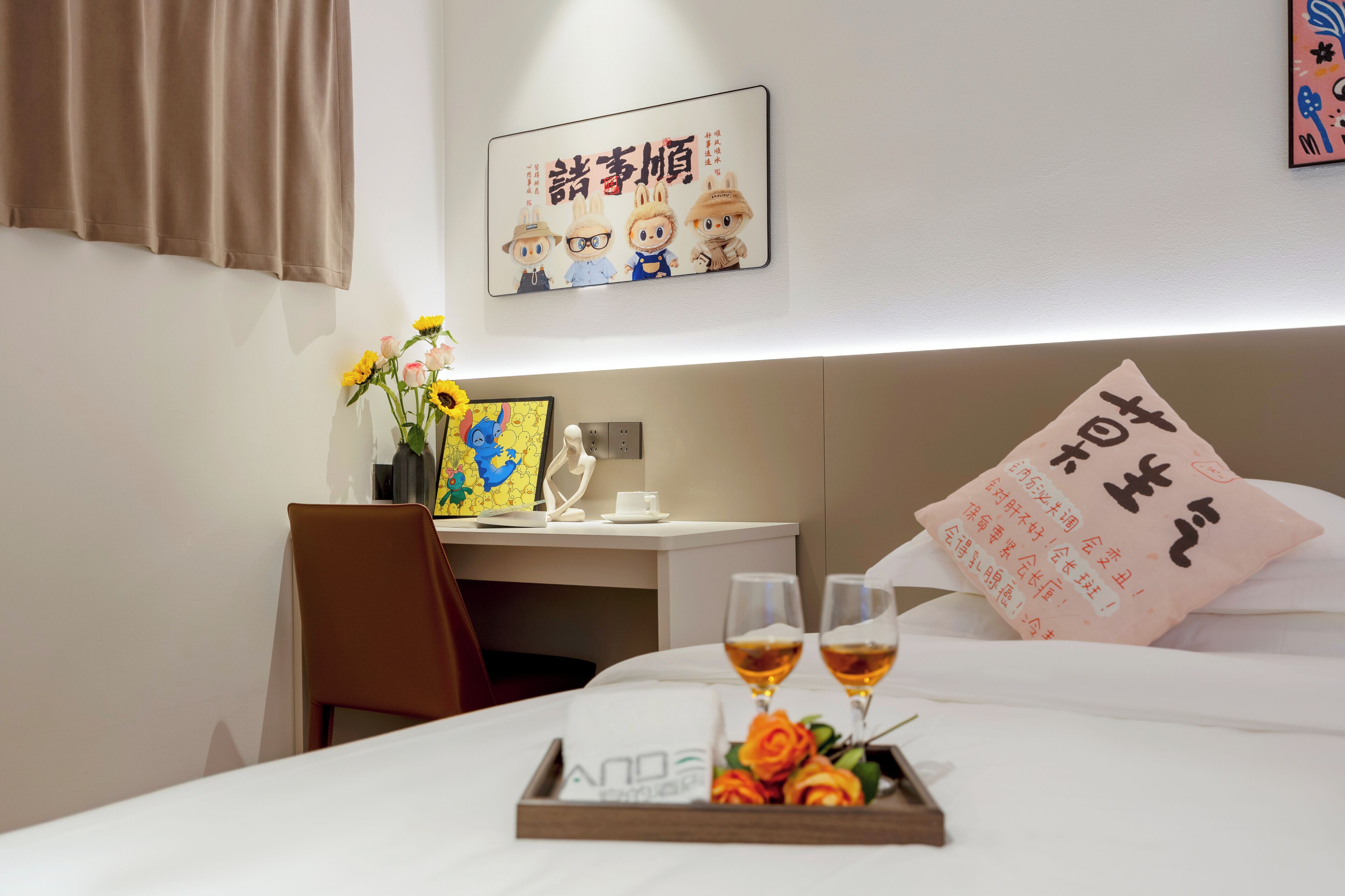 Quarto de hotel com cama bem arrumada, mesa de cabeceira com um vaso de flores, quadro na parede e almofada com texto.