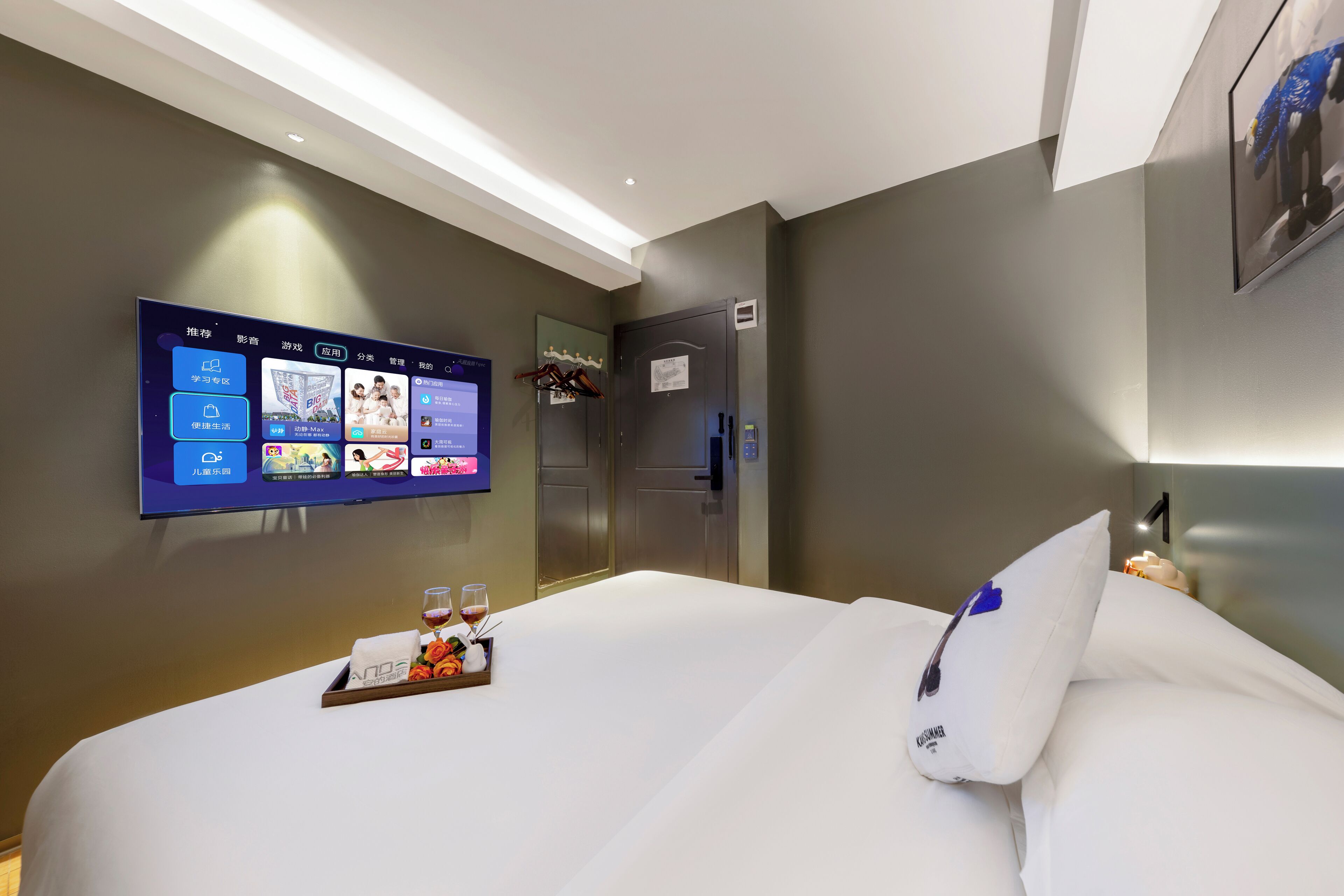 Habitación de hotel con un televisor de pantalla plana grande montado en la pared que muestra varias aplicaciones y un menú. Hay una cama con ropa blanca y un atril con aperitivos y una copa de vino.