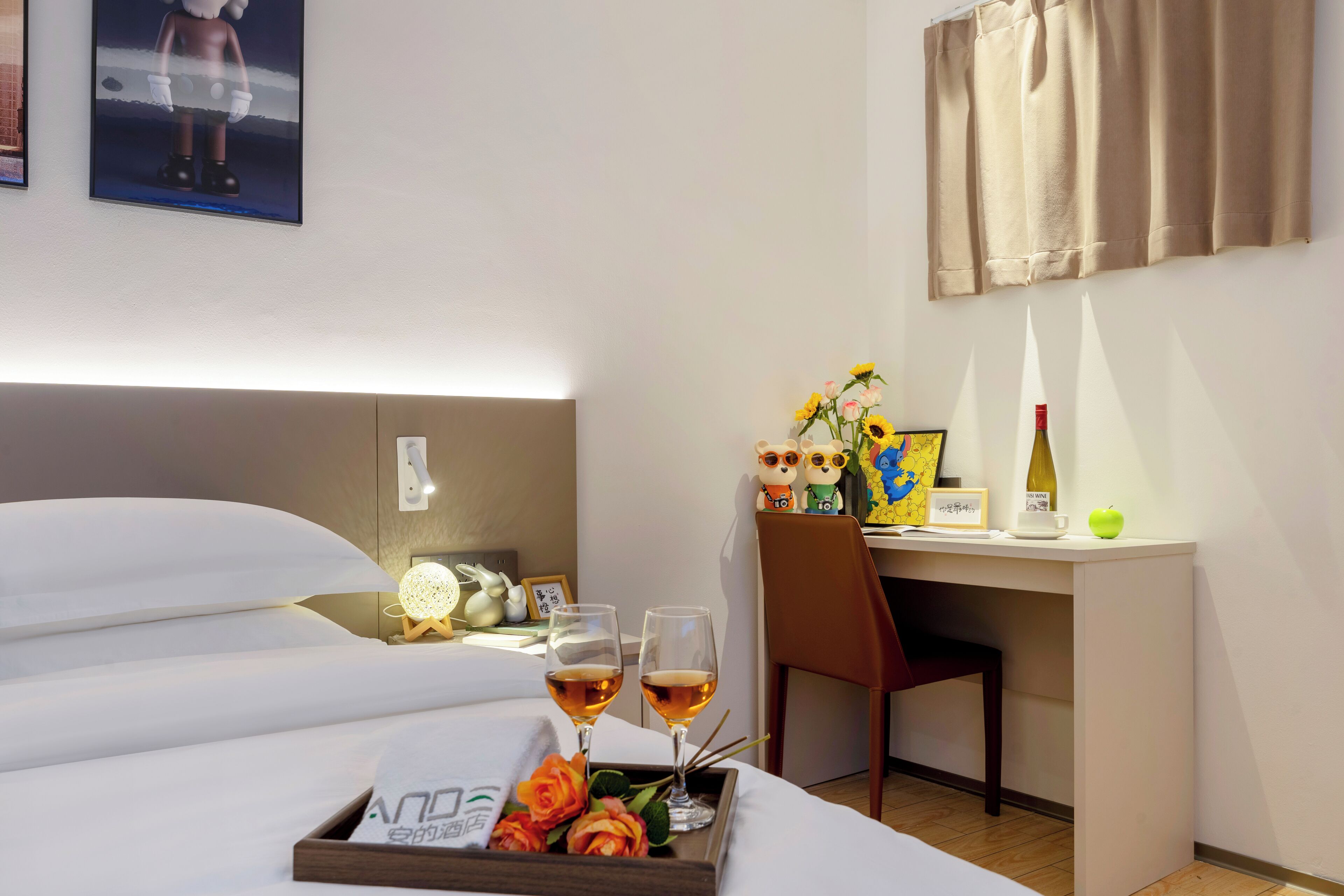 A hotel room with a bed, a desk with a chair, a tray with flowers and glasses of wine, and a wall with a picture and a bottle.