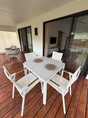 A covered outdoor patio with a white table and chairs, a TV mounted on the wall, and a sliding glass door leading to an interior space.