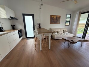 Una cocina moderna con armarios blancos, un comedor con mesa de madera y sillas blancas, un sofá con cojines decorativos, y un amplio ventanal con vistas a zonas verdes.
