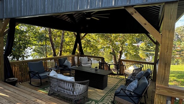 A covered outdoor deck with wooden flooring, a central seating area with a table and chairs, and a view of a lake surrounded by trees. - Large Pond View, 2 min to Lake! Hot tub (Nancy)