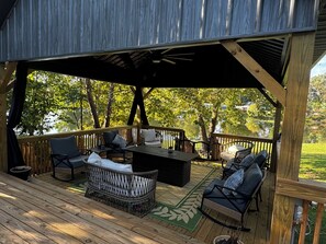 A covered outdoor deck with wooden flooring, a central seating area with a table and chairs, and a view of a lake surrounded by trees. - Large Pond View, 2 min to Lake! Hot tub (Nancy)