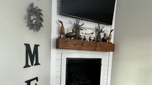 A living room with a fireplace, a mounted TV, and a wall-mounted shelf with decorative items. - Large Pond View, 2 min to Lake! Hot tub (Nancy)