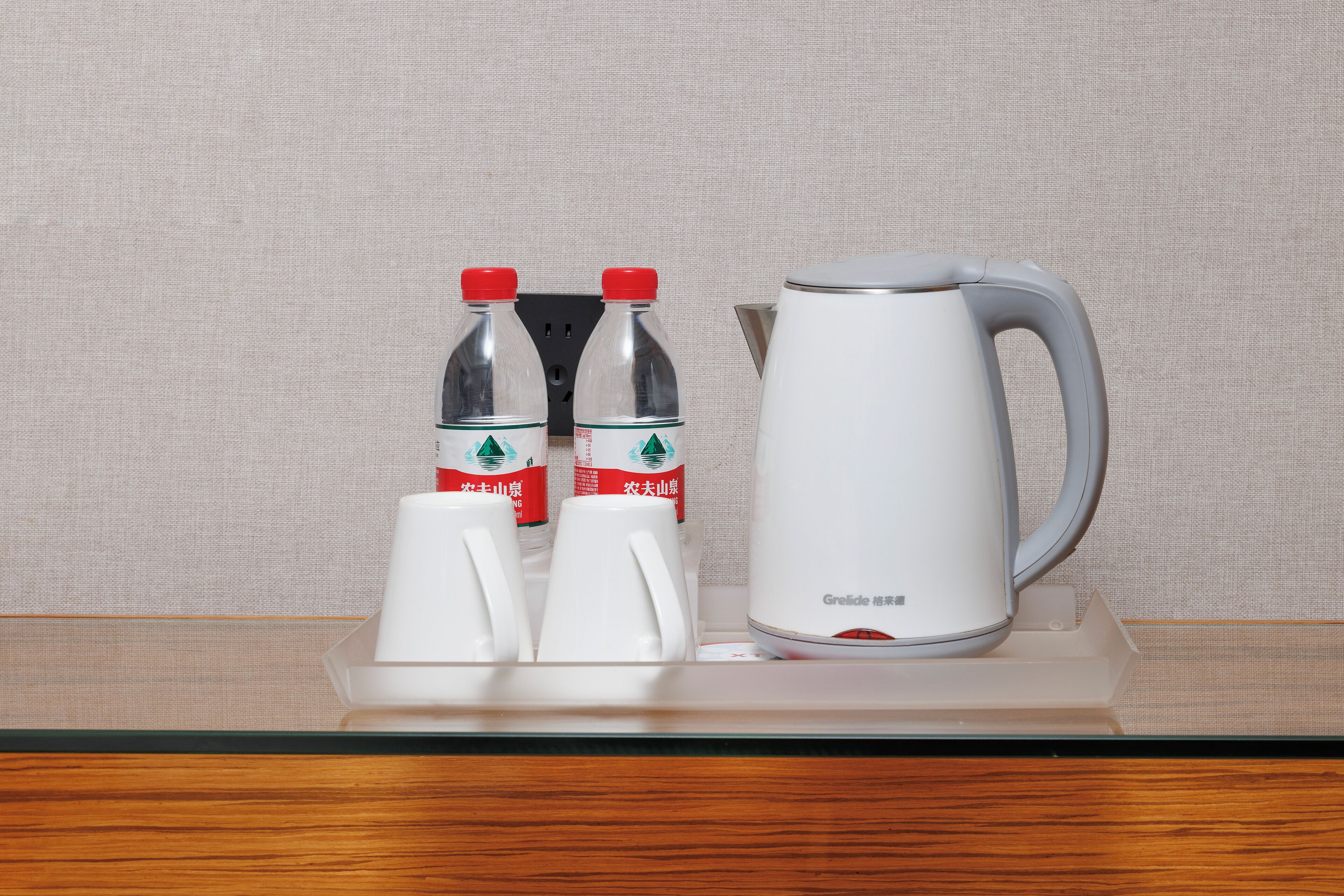 A hotel room's minibar with a white kettle, two bottles of water, and two cups.