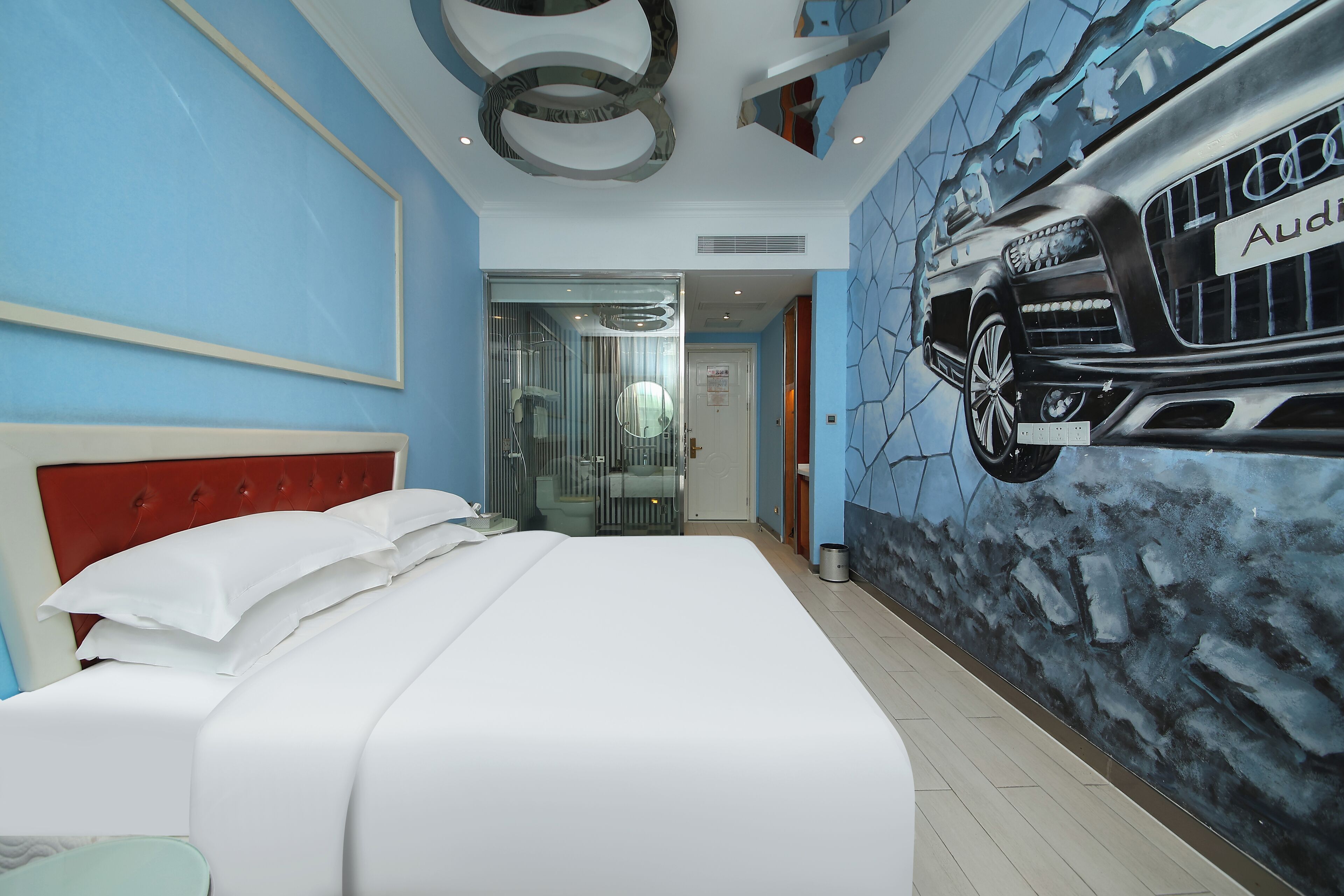 A hotel room with a bed, a red headboard, a wall mural of an Audi car, and a glass shower enclosure.