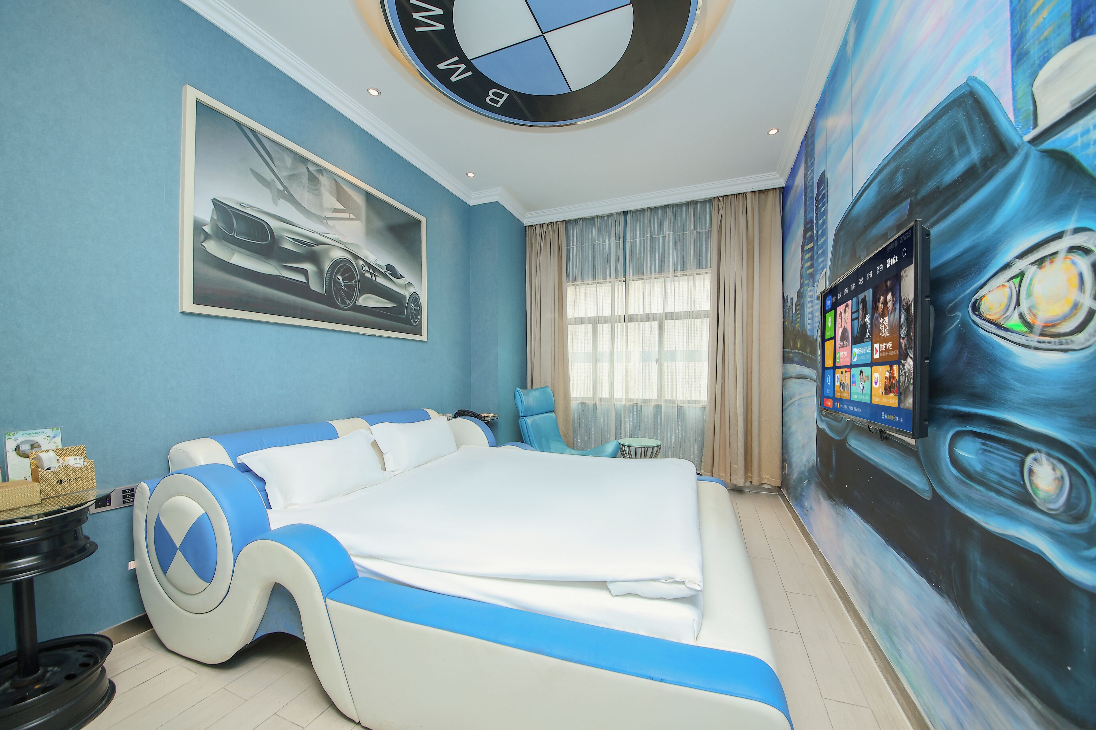 A modern hotel room with a large bed, a BMW logo on the ceiling, a cityscape mural, and a digital screen displaying content.