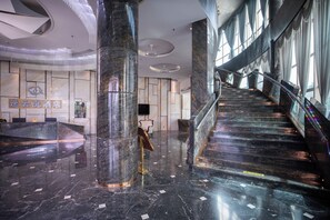 Un hall d’hôtel moderne avec un sol en marbre, un comptoir d’accueil et un escalier menant aux étages supérieurs.