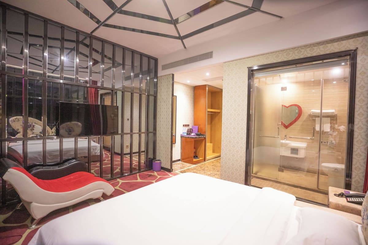 Ein Hotelzimmer mit einem Bett, einer Sitzecke mit einem rot-weißen Stuhl, einer Duschkabine aus Glas und einem kleinen Tisch mit einem Laptop.