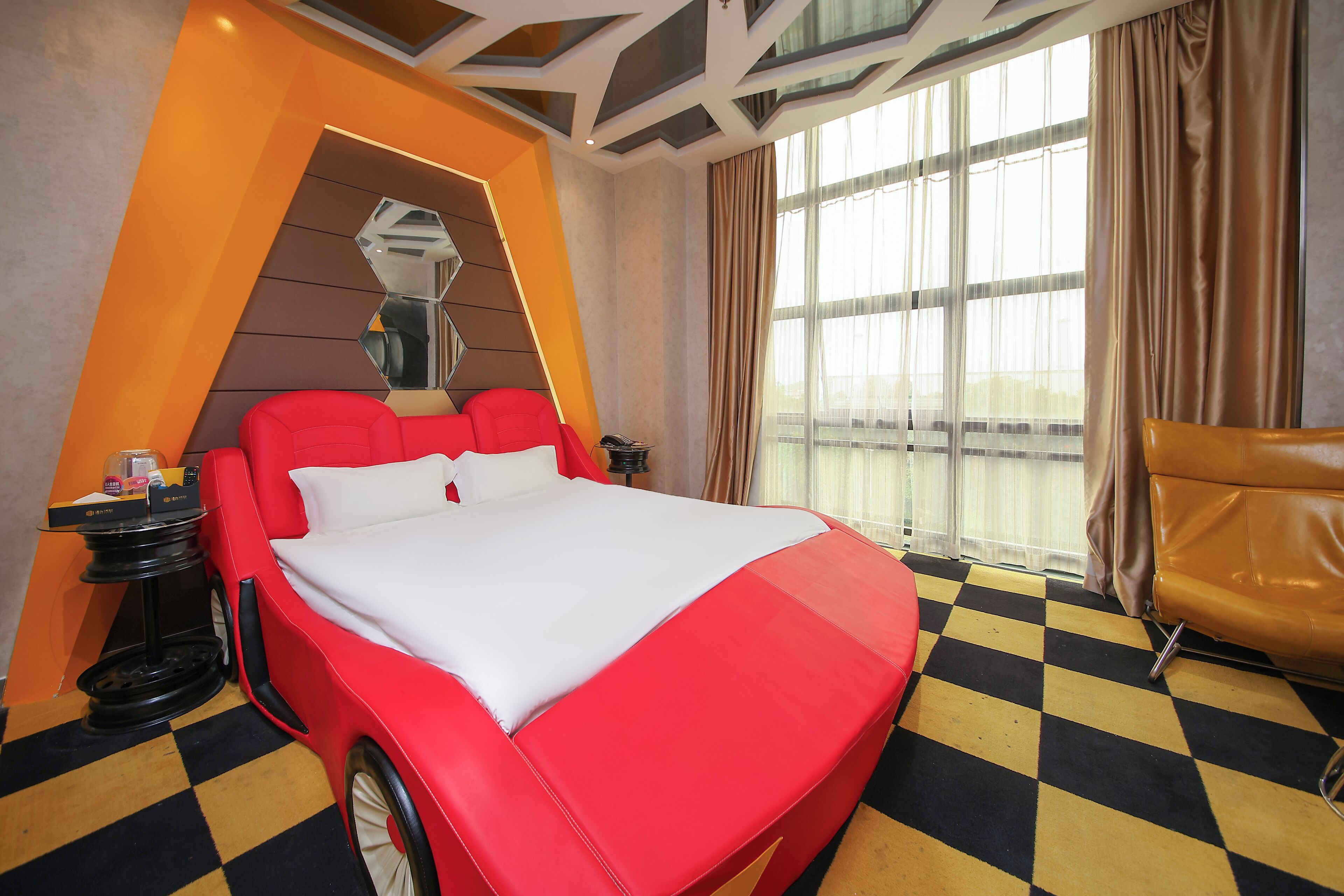 Une chambre d’hôtel moderne avec un grand lit, une tête de lit rouge, un sol à carreaux et une grande fenêtre avec des rideaux.
