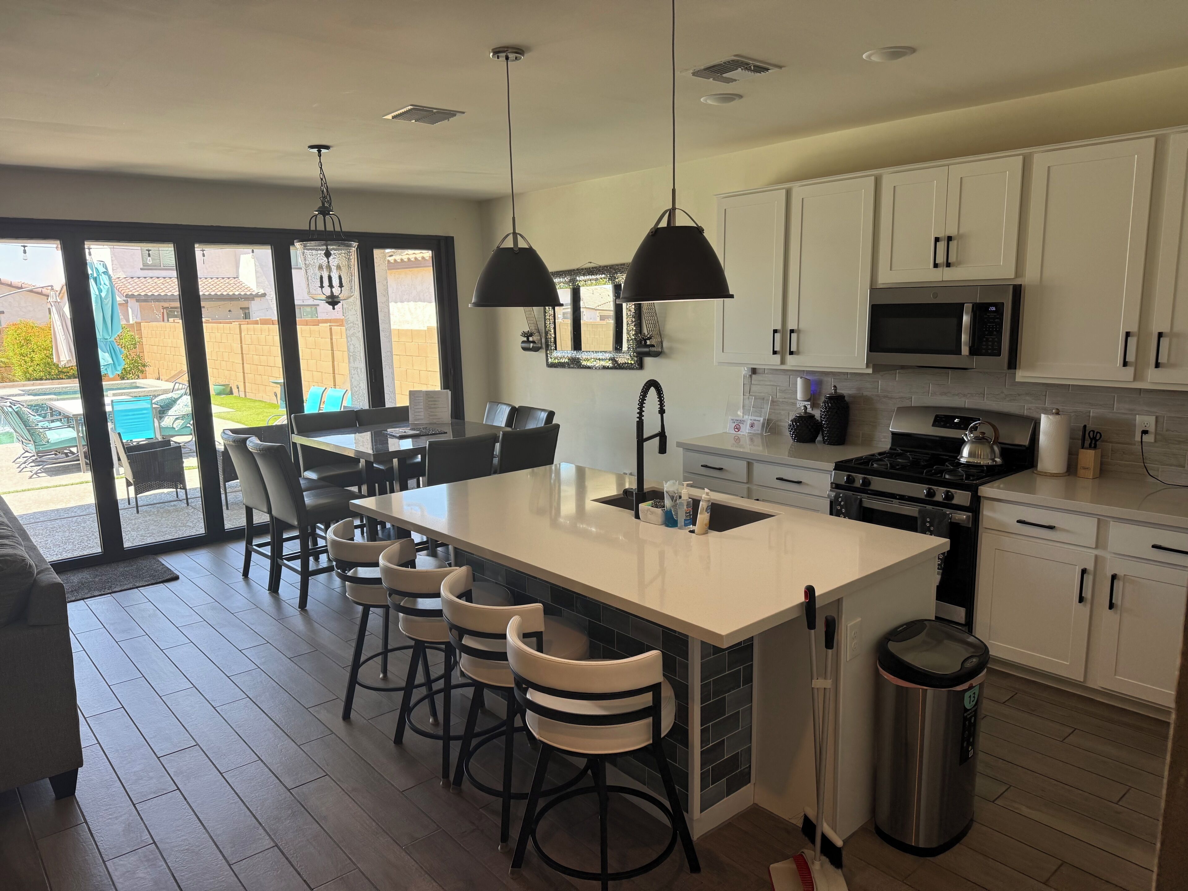 A modern kitchen with white cabinets, a central island with bar stools, a stainless steel stove, and a large window overlooking a backyard.