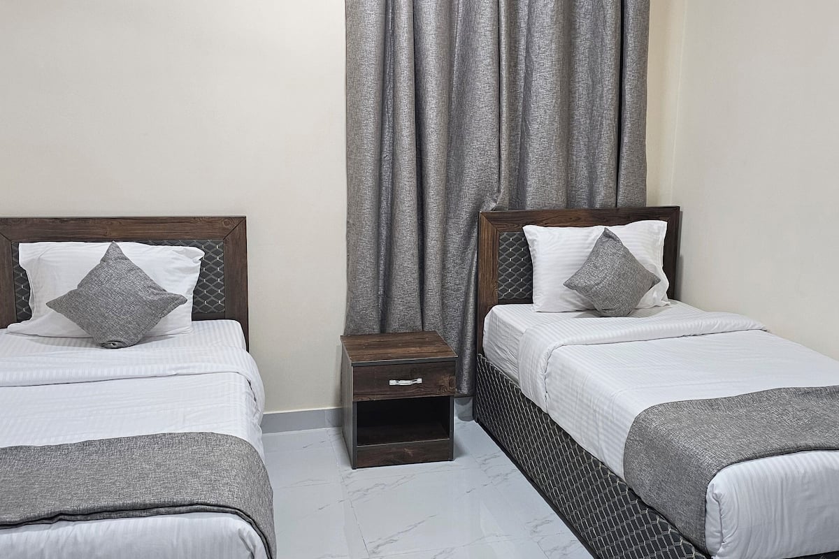 A hotel room with two single beds, a nightstand, a chair, and a window with grey curtains.