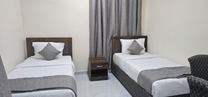 A hotel room with two single beds, a nightstand, a chair, and a window with grey curtains. - Today House Suites 3 (Salalah)