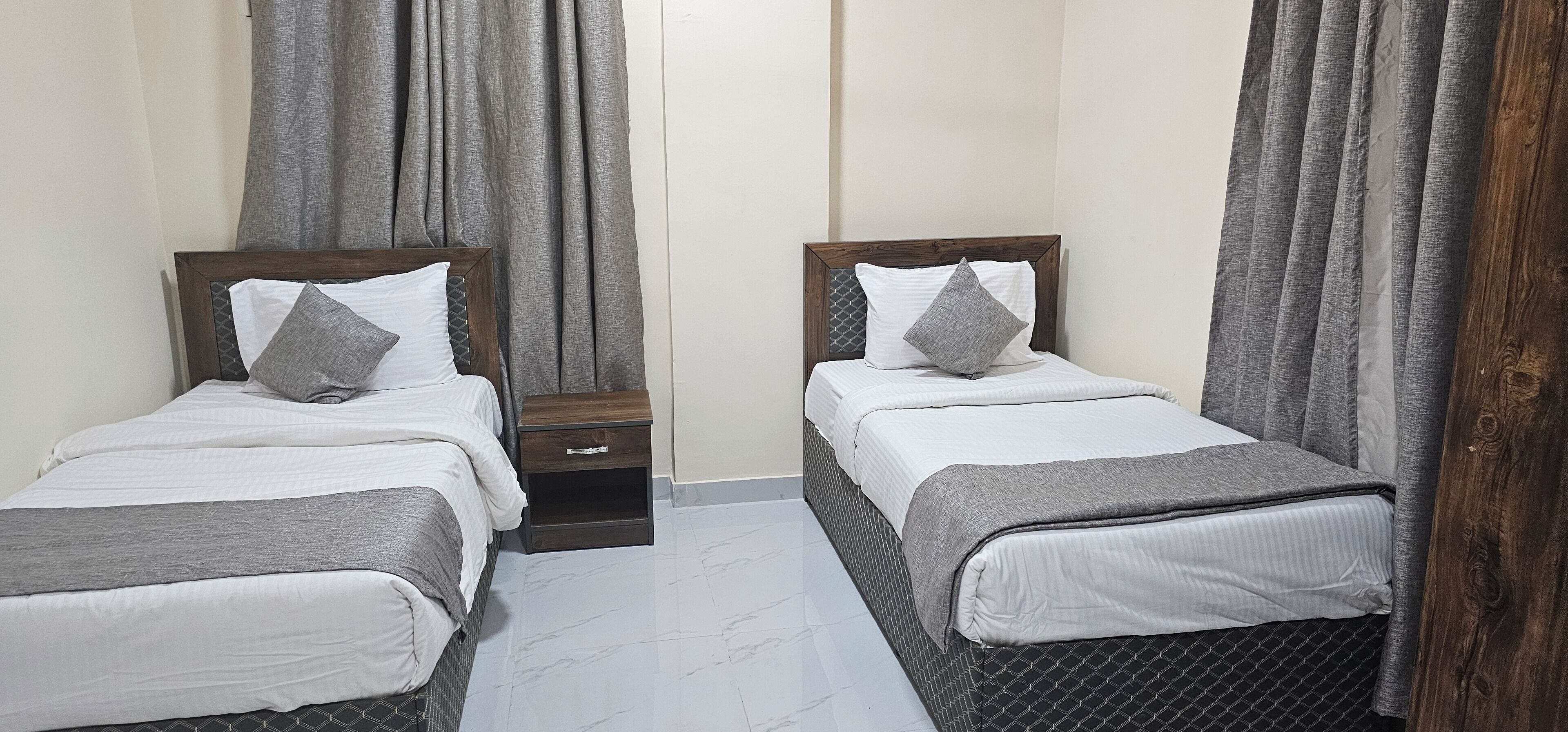 A twin room with two single beds, each with a wooden headboard, grey bedding, and a grey pillow. There is a small wooden nightstand with a drawer between the beds.