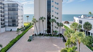 A modern hotel with a central courtyard, palm trees, and a view of the ocean. - Admiralty on Gulf Shore Dr. - 6th flr Beachfront Penthouse - Amazing Gulf views & Stunning Sunsets (Naples)