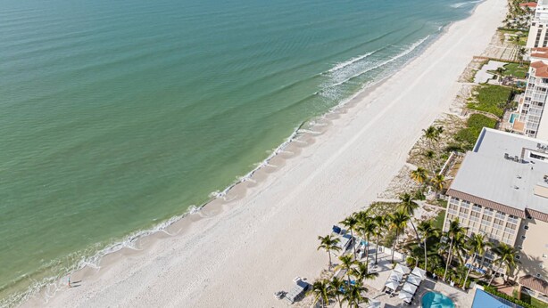 An aerial view of a beachside hotel with a long sandy beach, a pool, and several palm trees. - Admiralty on Gulf Shore Dr. - 6th flr Beachfront Penthouse - Amazing Gulf views & Stunning Sunsets (Naples)