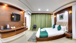 A modern hotel room with a curved wooden desk, a flat-screen TV, a bed with green bedding, a chair, and a small table with a phone and a plant. - HOTEL GK PALACE (Guwahati)