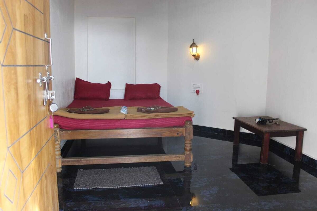A room with a wooden bed, red bedding, a small wooden table, and a wall-mounted light fixture.