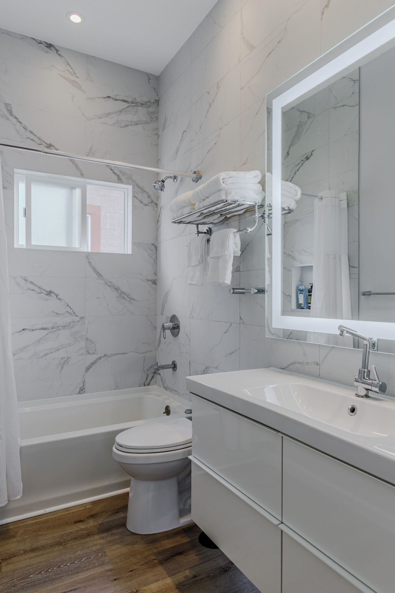 A modern bathroom with a marble wall, a bathtub, a toilet, and a vanity with a mirror.