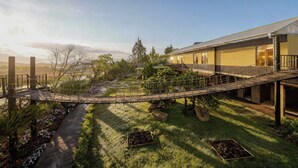Un puente de madera que conecta dos edificios en un jardín ajardinado, con vistas a colinas distantes.