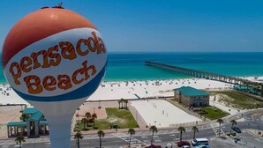 En strandscene med et stort skilt, der viser "Pensacola Beach", og en mole, der strækker sig ud i vandet.
