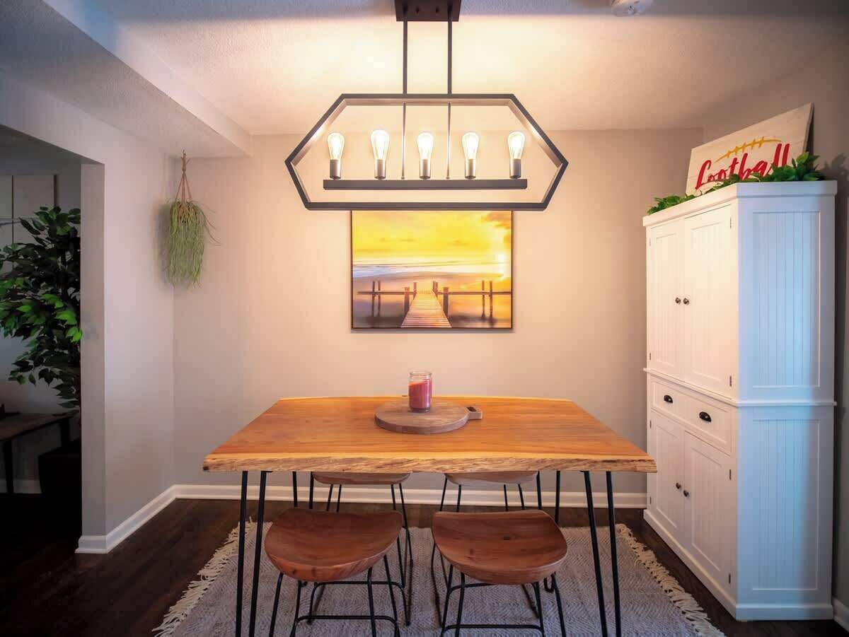 A dining area with a wooden table, two chairs, a hanging light fixture, a painting, and a cabinet.