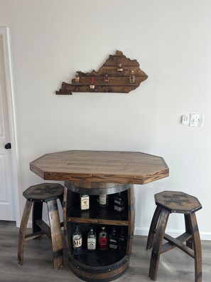 Une table ronde en bois, accompagnée de deux tabourets, une étagère en bois fixée au mur avec des bouteilles, et une porte blanche en arrière-plan.