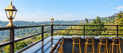 A terrace with a glass balustrade, a row of bar stools, and two street lamps.